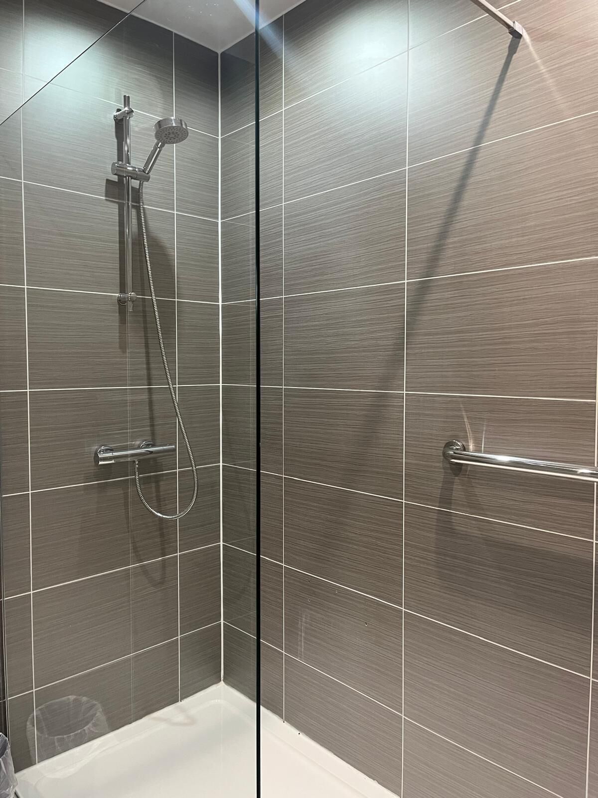 Una ducha moderna con cabezal de ducha manual y grifo empotrado en la pared, zona de ducha con mampara de vidrio y una bañera blanca.