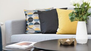 A modern living room with a grey sofa, three patterned cushions, a white vase with a plant, and a small round table with a magazine and a geometric object. - The Briarcroft Wayth Hotels (Goole)
