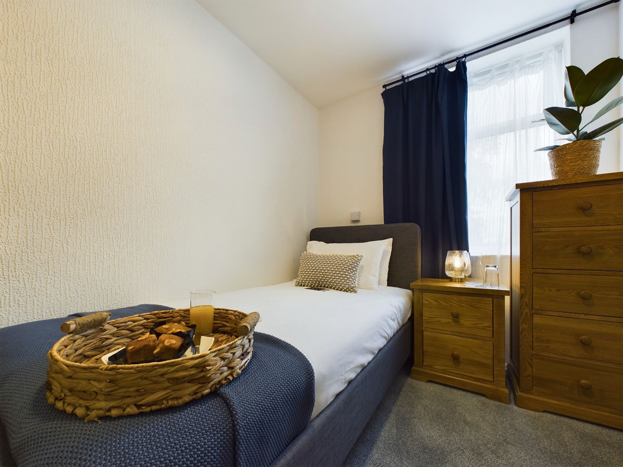 Un dormitorio ordenado con una cama, un tocador de madera, una mesita de noche con lámpara y un ventanal con cortinas.