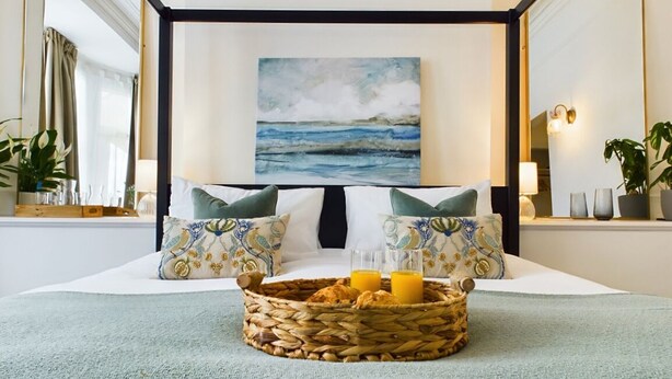 A hotel room with a bed, a painting of the sea, a bedside table with glasses and a basket of pastries, and a large mirror. - The Briarcroft Wayth Hotels (Goole)