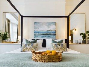 A hotel room with a bed, a painting of the sea, a bedside table with glasses and a basket of pastries, and a large mirror. - The Briarcroft Wayth Hotels (Goole)