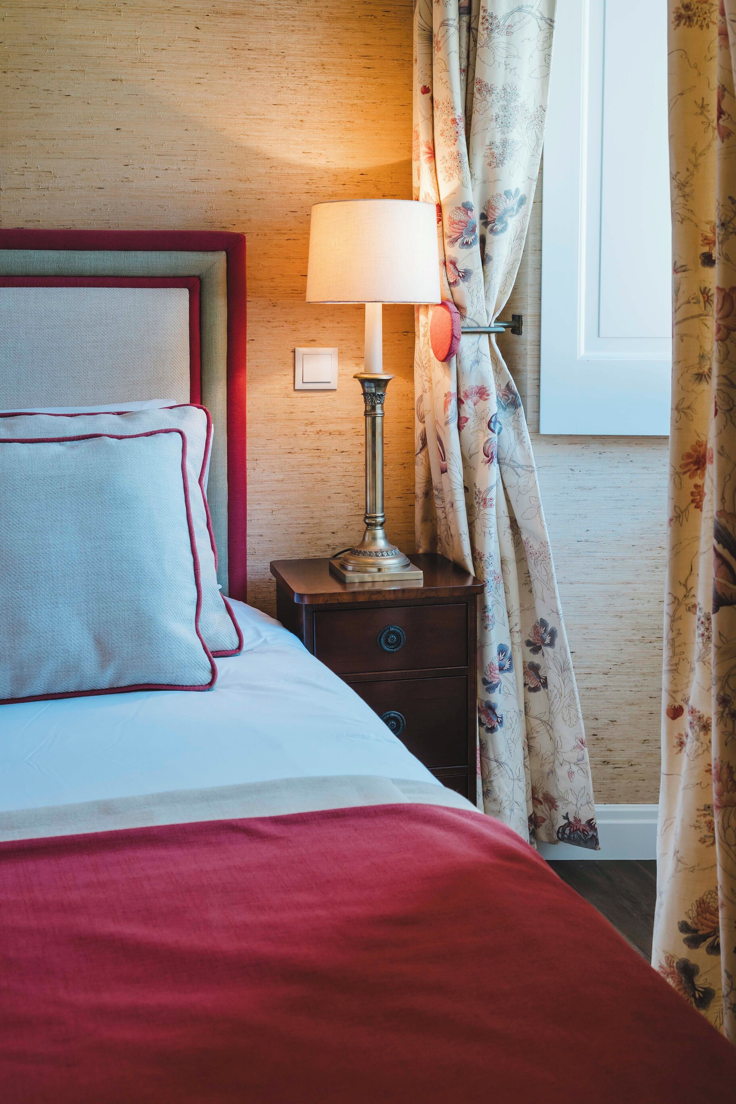 Una cama bien hecha con una manta y almohadas rojas, una mesita de noche con una lámpara y cortinas con estampado floral.