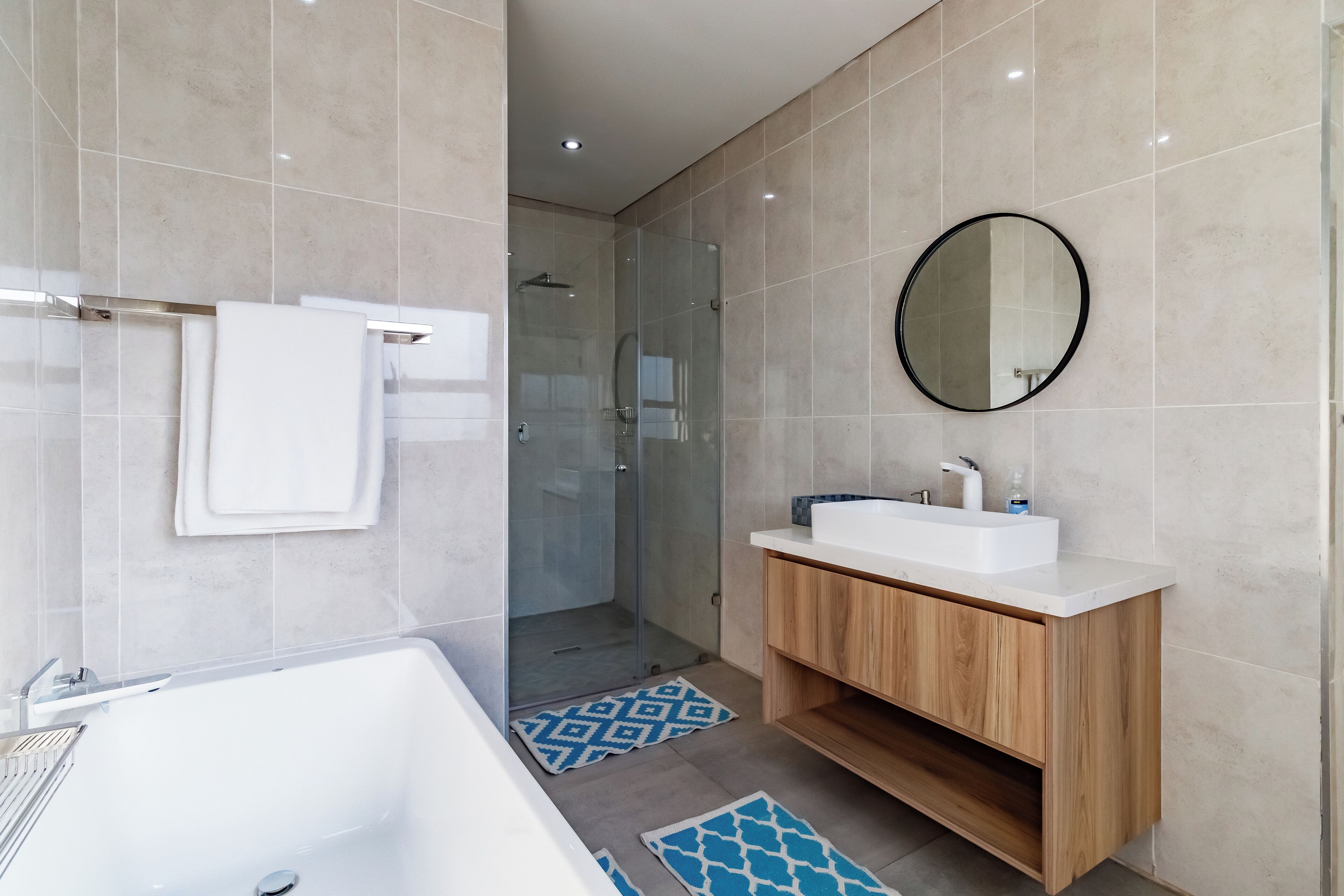 A modern bathroom with a bathtub, a glass-enclosed shower, a wooden vanity with a white sink, and a round mirror.