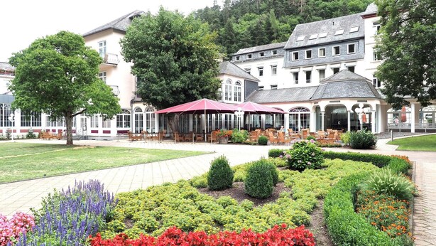 A hotel with a garden patio area, outdoor seating, and a large building with multiple windows and a covered section. - Holiday Apartment Wasserfall in Holiday Home Bad Bertrich (Bad Bertrich)