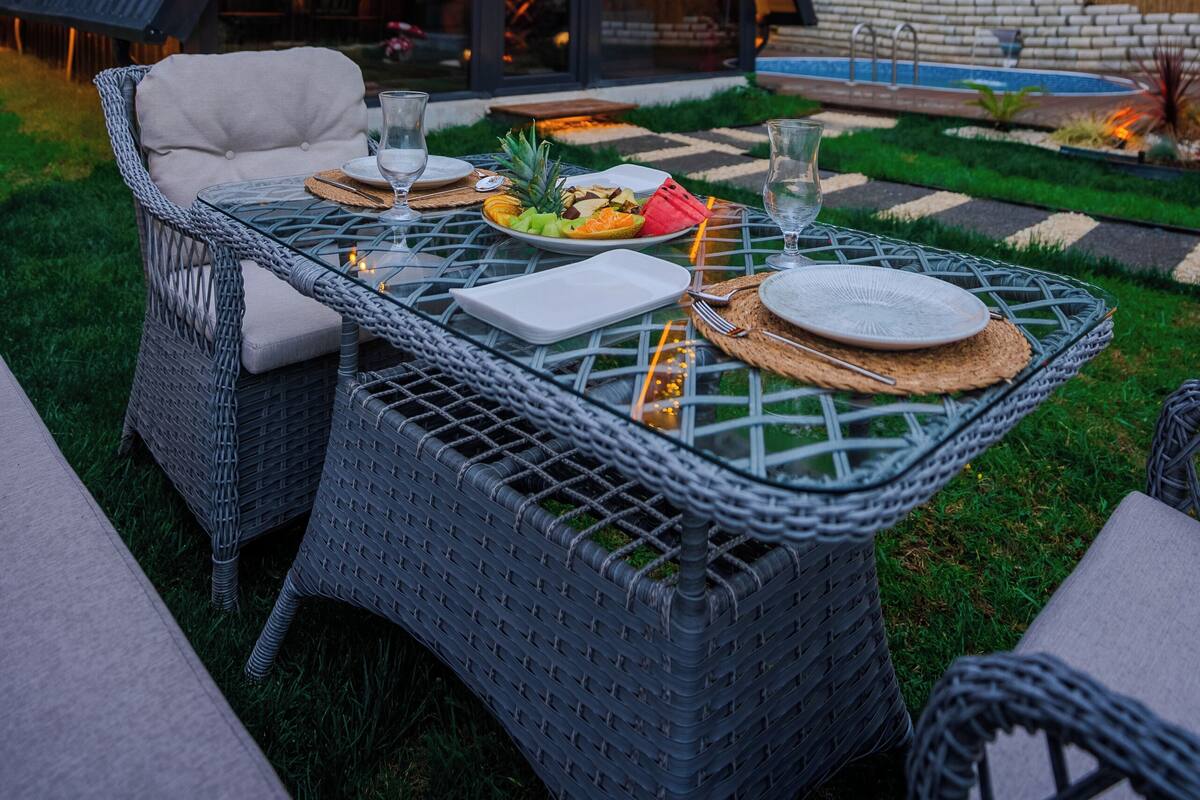 A modern outdoor dining area with wicker furniture, a glass table, and a view of a building with a triangular roof and string lights.