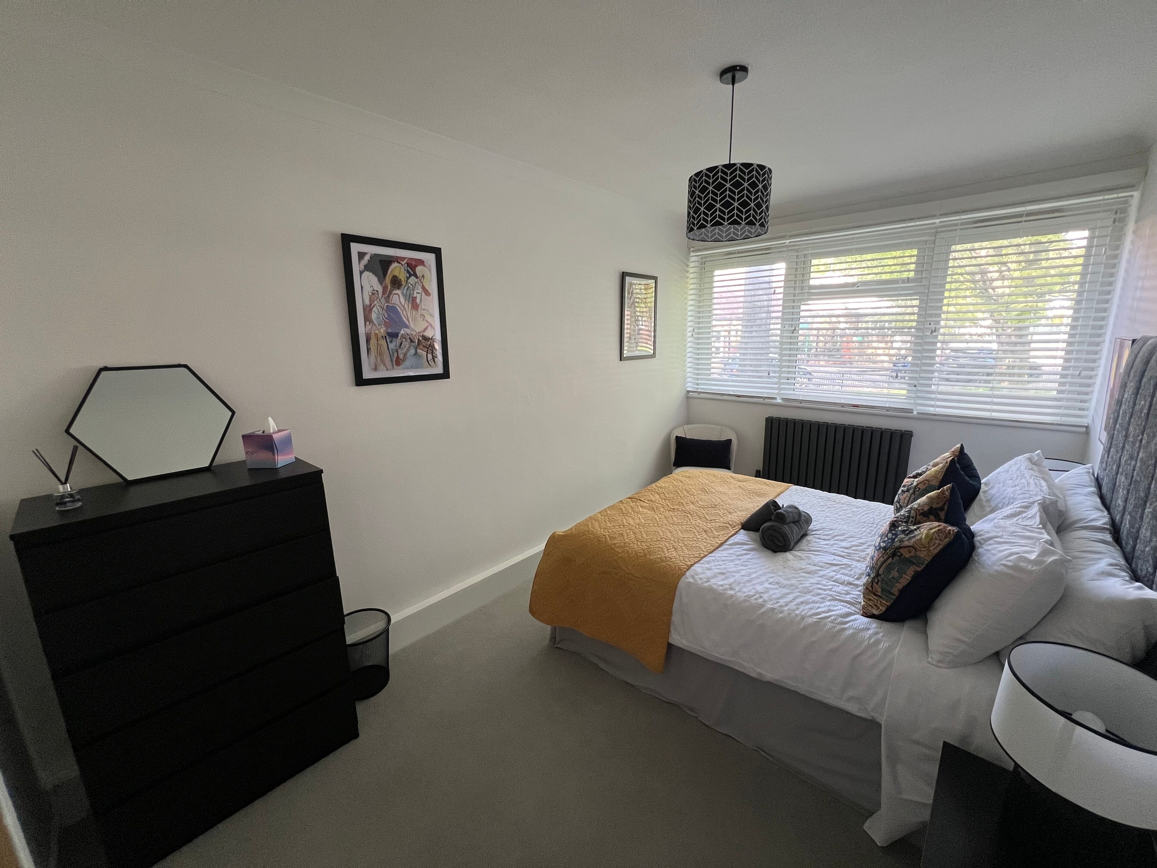 Une chambre à coucher comprenant un lit, une commode, une chaise, une fenêtre avec des stores et un luminaire suspendu.