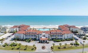 Um hotel à beira-mar com vários edifícios, uma piscina central e estacionamento.