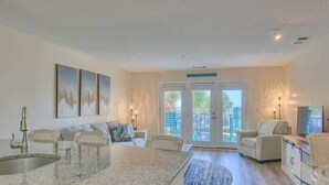 A spacious living area with a kitchen, dining table, and seating. There are large windows and a sliding glass door leading to an outdoor area. - Villa Capriani 102B - Oceanfront - Pools - Restaurant - Premiere (North Topsail Beach)