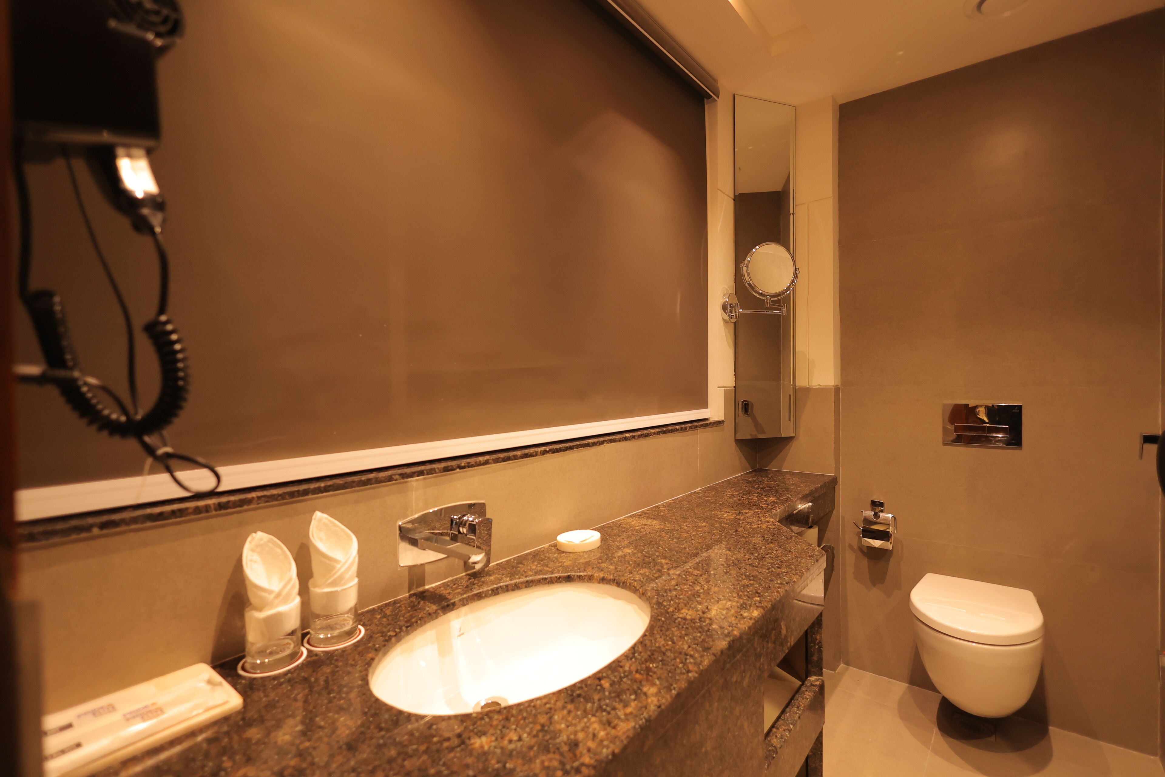 A bathroom with a granite countertop, a sink, a mirror, a toilet, and a hairdryer on the wall.