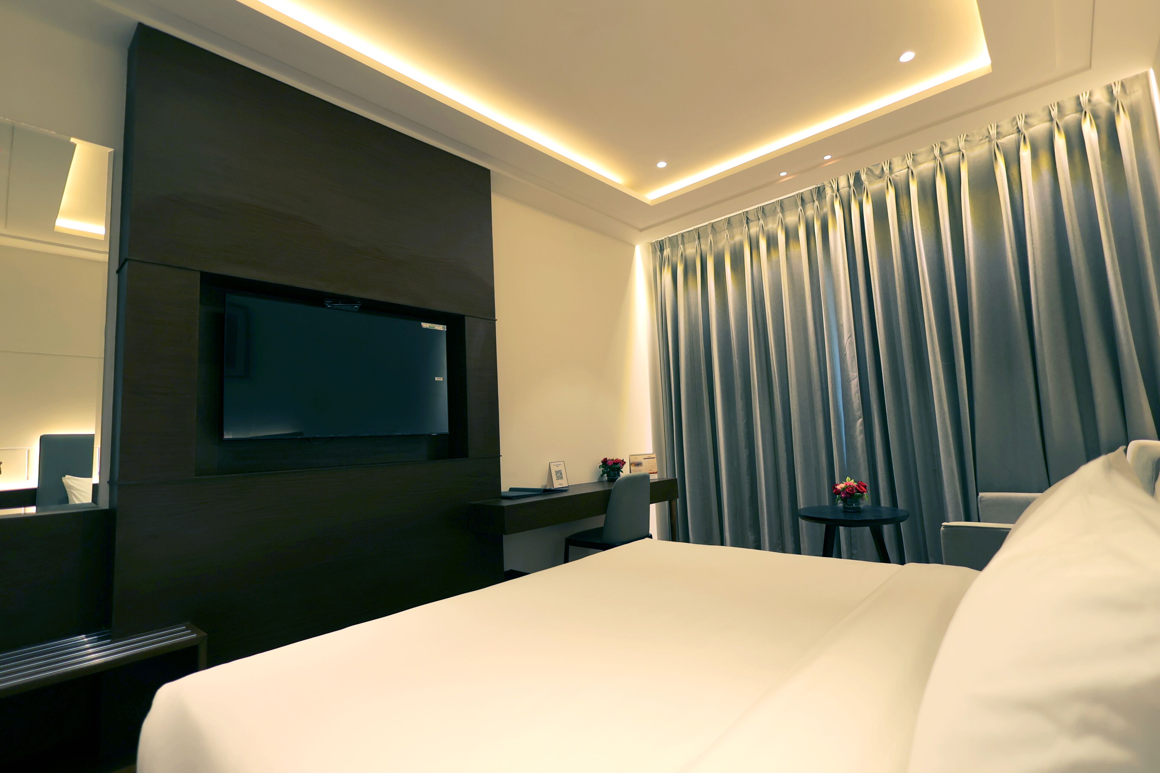 A modern hotel room with a large bed, a desk, a flat-screen TV mounted on the wall, and a window with curtains.