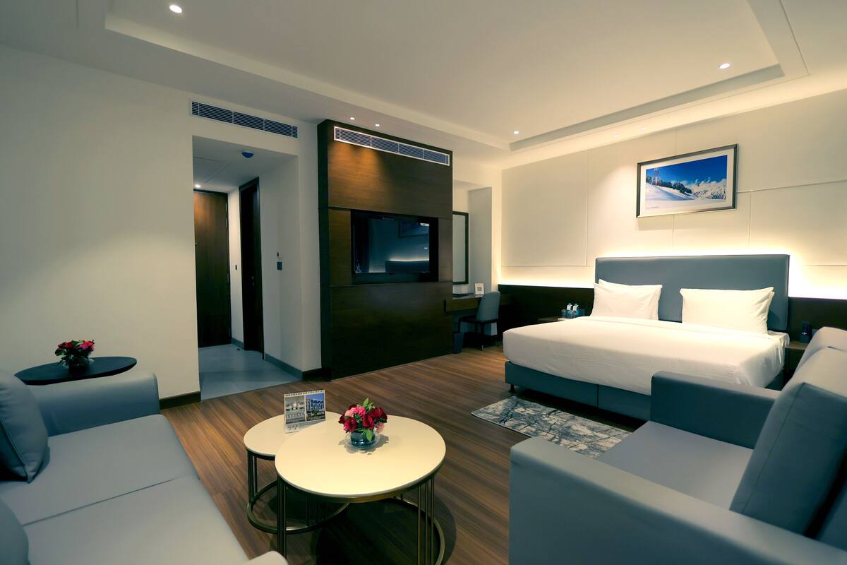 Ein modernes Hotelzimmer mit einem großen Bett, einem Fernseher, einem Schreibtisch und einer Sitzecke mit einem kleinen Tisch und Stühlen.