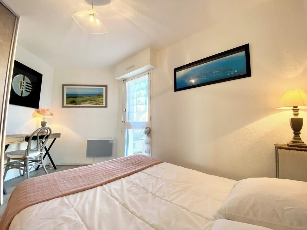 Een slaapkamer met een bed, een bureau en een stoel. Er is een raam met gordijnen, een schilderij aan de muur en een lamp op het nachtkastje.
