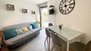 A living room with a sofa, a dining table with chairs, a large wall clock, and a small plant on the table.