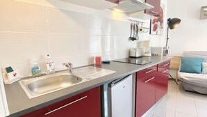 A modern kitchen with red cabinets, a stainless steel sink, and a dishwasher. There is a window with blinds, a coffee maker, and a lamp on the counter.