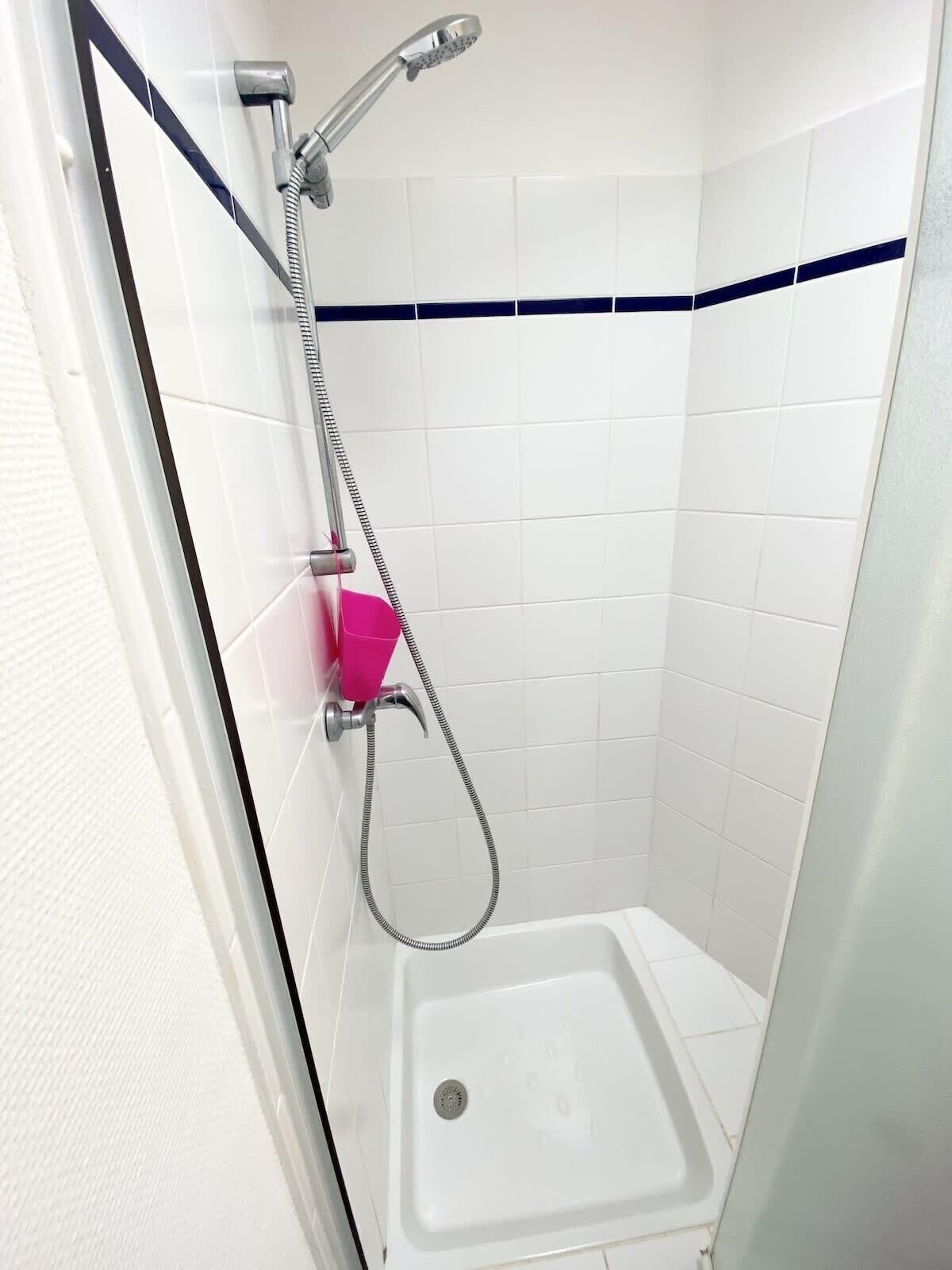A small, white-tiled shower room with a single showerhead and a pink soap dispenser.