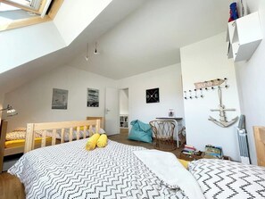 A bedroom with a loft-style ceiling, a bed with patterned bedding, a wooden headboard, a small table with books, a chair, and a wall-mounted shelf with decorative items.