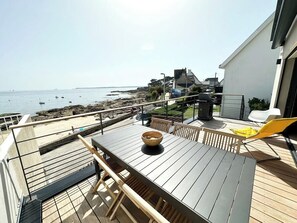 Eine Holzterrasse mit Tisch und Stühlen, mit Blick auf einen Strand, im Hintergrund Boote und Gebäude.
