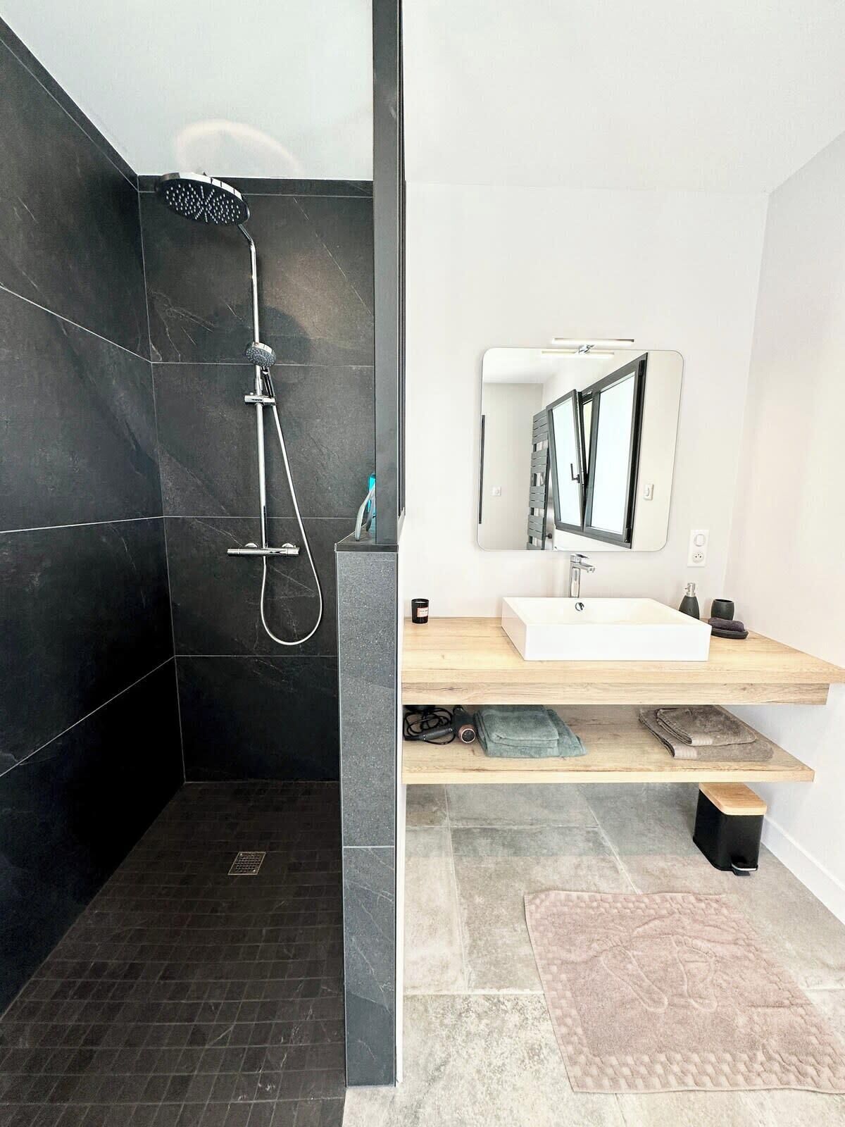 A modern bathroom with a dark tiled shower, a white sink with a wooden countertop, and a large mirror.