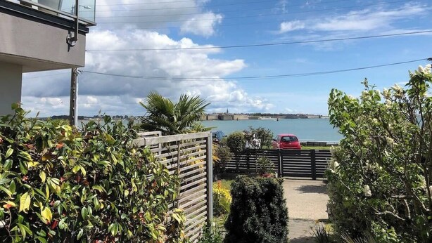 Un quartier résidentiel avec vue sur une étendue d’eau, une voiture rouge garée près d’une porte, et un immeuble doté d’un balcon surplombant le paysage.