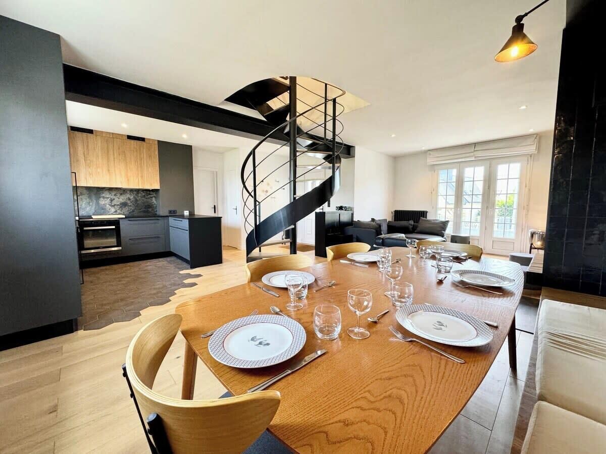 Un comedor moderno con una mesa de madera dispuesta para seis personas, una escalera en espiral y una cocina al fondo.