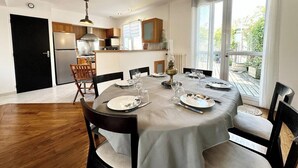 A dining area with a round table set for six, surrounded by wooden chairs. There is a kitchen in the background with modern appliances and wooden cabinets.