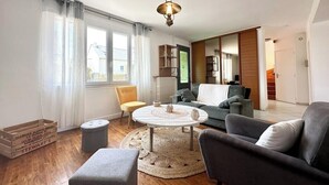 A modern living room with a round coffee table, sofas, and a yellow chair. There is a wooden floor, a large window with curtains, and a staircase in the background.