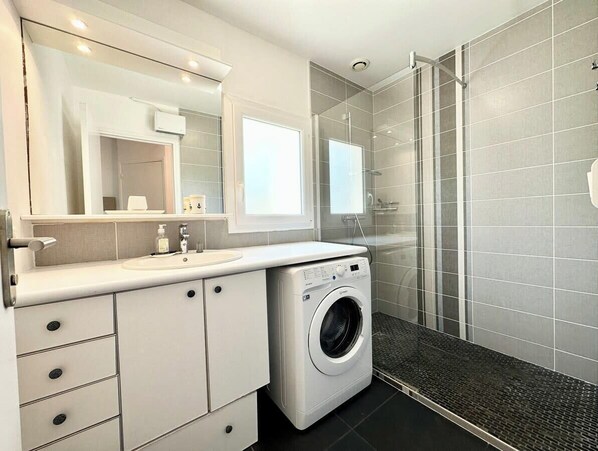Un baño moderno con lavadora y secadora integradas, cerramiento de ducha de vidrio y un espejo grande sobre el lavabo.