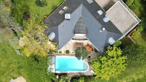 An aerial view of a house with a swimming pool, surrounded by greenery and a paved driveway.