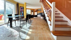 A spacious living area with a glass dining table, comfortable seating, a wooden staircase, and a large window offering a view.