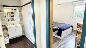 Una habitación con cama, escritorio y ventana. Cuenta con un baño con lavamanos y espejo. El piso es de baldosas y tiene una lámpara en el techo.