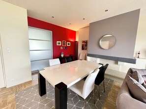 A modern dining area with a white table and black chairs, a red accent wall, and a large mirror.