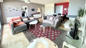 A modern living room with grey sofas, a coffee table, and a TV on a glass table. There is a patterned rug on the floor, and a red wall with a window. - Luxury T3 standing 2 - Vue mer exceptionnelle 180° (Larmor-Plage)