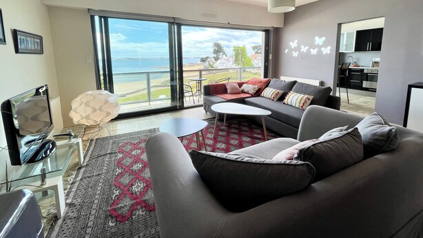 A modern living room with a grey sofa, a glass coffee table, and a patterned rug. There is a large window offering a view of the beach and ocean. - Luxury T3 standing 2 - Vue mer exceptionnelle 180° (Larmor-Plage)