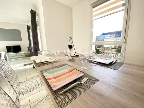 A modern dining area with a long table, clear glass chairs, and a television in the background.