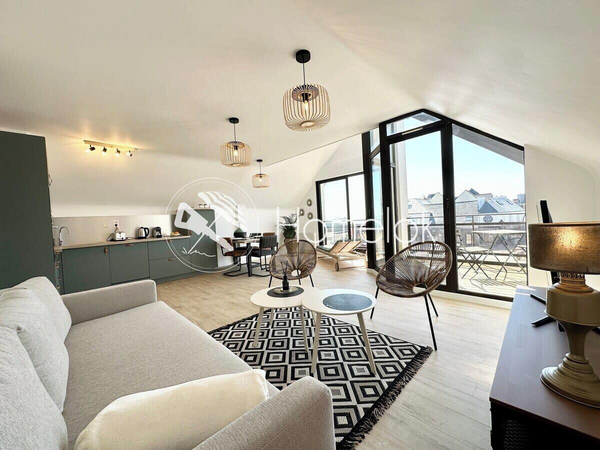 Een moderne woonkamer met een bank, een eettafel en stoelen. Er is een keuken met kasten en een spoelbak. De kamer heeft grote ramen met uitzicht op gebouwen.