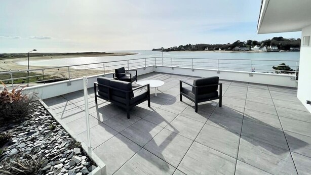 A rooftop terrace with two black sofas and a small round table, overlooking a beach and the sea.