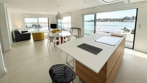 A modern living room with a white sofa, yellow ottoman, and a dining area with a round table and chairs. There is a large window with a view of the sea.
