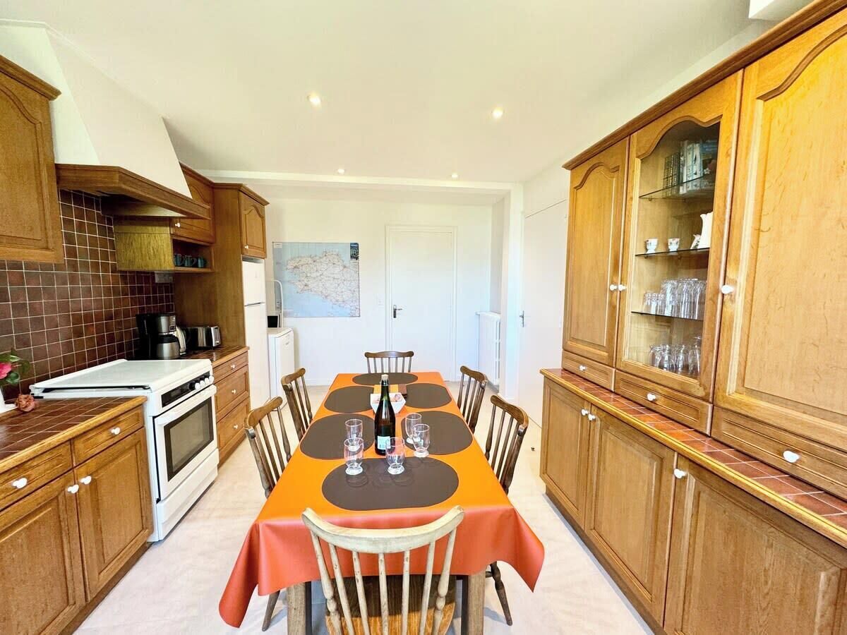 Une cuisine avec des meubles de rangement en bois, une table à manger recouverte d’une nappe rouge et un réfrigérateur.