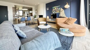 Un salon moderne avec un canapé, un fauteuil et une table basse. On y trouve également un coin repas avec des chaises et une table. Un téléviseur est fixé au mur.