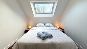 Un dormitorio con tragaluz, cama con ropa de cama blanca, dos almohadas y dos lámparas de noche.
