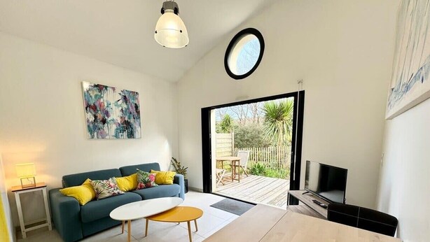 Una sala moderna con sofá, mesa de centro y televisor. Cuenta con un balcón con piso de madera y vista al jardín.