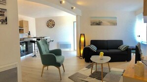 Een moderne woonkamer met een zwarte bank, een groene fauteuil en een kleine ronde tafel. Achtergrond toont een keuken met een klok aan de muur.