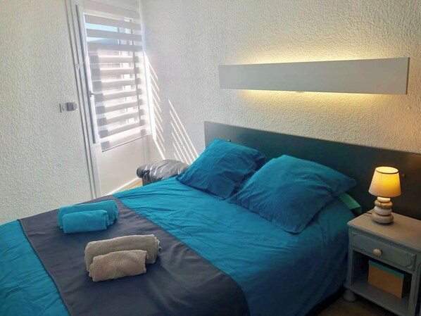 A bedroom with a bed, pillows, a nightstand, a lamp, and a window with blinds.