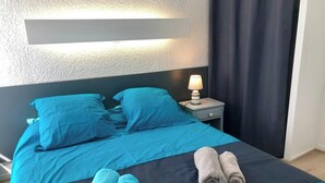 Een opgemaakt bed met blauw beddengoed en twee handdoeken, een bedlamp en een donker gordijn.