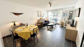 A spacious living area with a dining table set for four, a sofa, a coffee table, and a bookshelf. - Charmant T2 Top Cosy Face à la rade (Larmor-Plage)