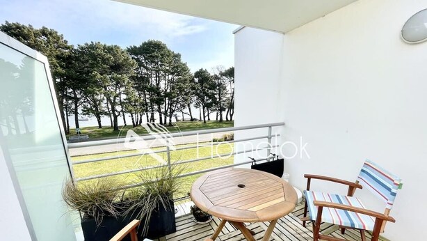 A balcony with a round wooden table and two striped chairs, overlooking a grassy area with trees. - Charmant T2 Top Cosy Face à la rade (Larmor-Plage)