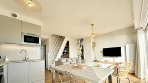 Una cucina moderna con forno a microonde integrato, un'area pranzo con tavolo in legno e sedie trasparenti, e un soggiorno con televisore e piante decorative.