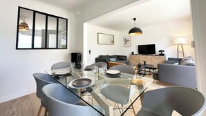 A modern living room with a glass dining table, grey chairs, a grey sofa, a TV, and a lamp.