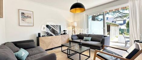 A modern living room with a sofa, a coffee table, and a chair. There is a TV on a stand and a window with curtains.