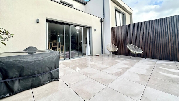 Une terrasse moderne dotée d’un barbecue couvert, de deux chaises blanches et d’une porte-fenêtre coulissante donnant sur une pièce entièrement vitrée.